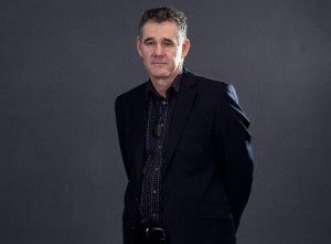 Photo of Paul Gibson.  He is pākehā and has short brown hair.  He is wearing a blue suit and shirt.  His taonga / pounamu is around his neck.